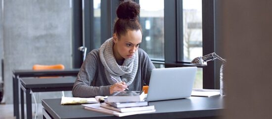 Student taking notes beside a laptop at a sunlit desk